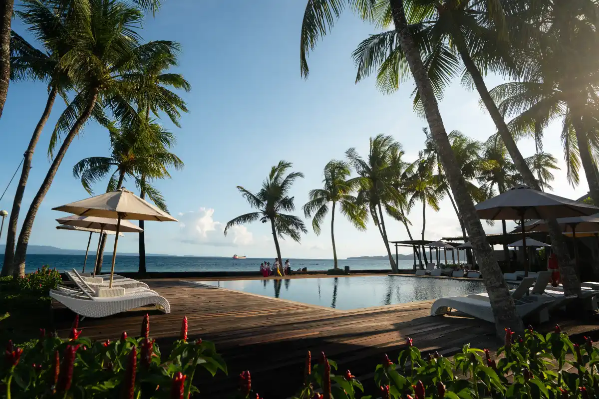 Nirwana Beach & Resort Candidasa outdoor swimming pool, wooden sunbathing deck, lounge chairs, and palm trees with ocean view.