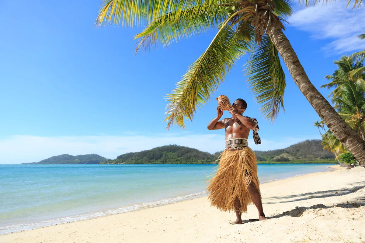 Fijian staff member performing traditional conch shell blowing welcome ceremony on Tropica Island Resort beach.