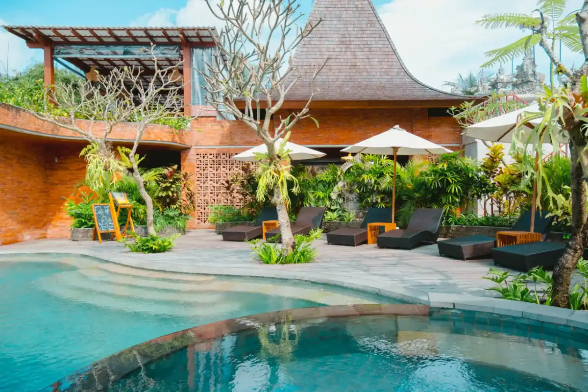 Outdoor swimming pool area at Emana Akatara with lounge chairs, sun umbrellas, and Balinese brick architecture.