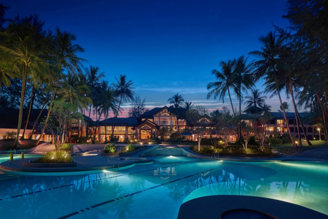 Outdoor swimming pool illuminated at dusk surrounded by palm trees and resort architecture at Dusit Thani Laguna Phuket.